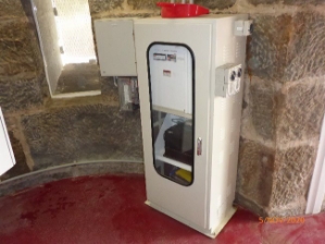 Photograph showing white electrical cabinet atop a red floor against a curved stone wall.