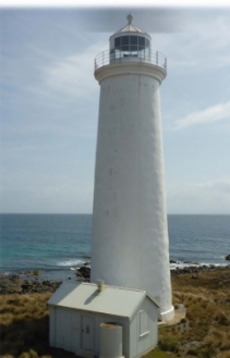 Photograph showing a smooth, white lighthouse tower beside a coastline, with a small white hut at its base.