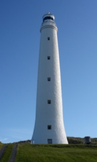 Photograph showing tall, white lighthouse tower with five windows running the length of the tower walls. Lighthouse is against a blue sky and sits on a green landscape. 