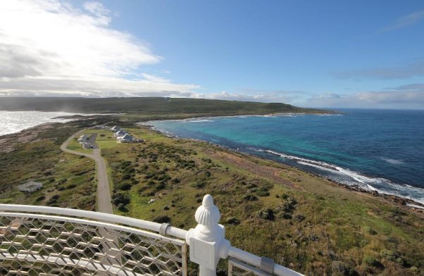 A photograph showing a collection of cottages on a green landscape surrounded by blue ocean. White balustrade seen in bottom-left corner.