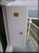Photographs showing white lantern room door standing ajar. Door opens onto narrow balcony with curved, white balustrades pictured behind.