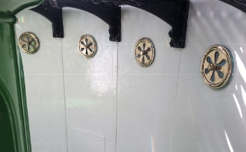 Photograph showing white wall panels. Four round, brass vents and black backets embedded in wall panels.