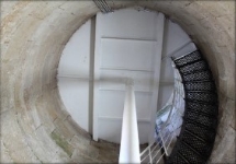 Photograph showing internal face of round, stone room. White underside of round floor above supported by while pole and floor beams. Black spiral stairs leading up to the white floor.