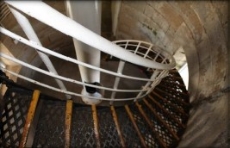 Photograph showing black cast iron staircase wrapping around stone walls, leading down to floor below. 