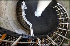 Photograph showing round floor with black spiral staircase leading to and away from level. White tube fixed to centre of floor. 