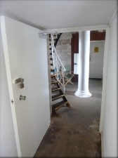 Photograph showing large door standing ajar, room through doorway has a large, white tube fitted in centre, and a spiral staircase leading from floor to above level.