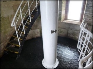 Photograph showing black, round iron floor. Curved stone wall encases floor, with white tube fitted to centre of floor. Black spiral stairs leading to floor above.