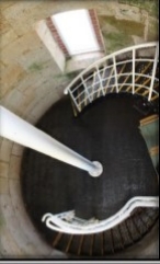 Photograph showing round floor with black spiral staircase leading to and away from level. White tube fixed to centre of floor. 