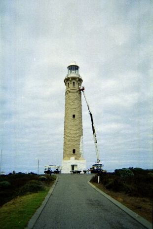 Photograph showing stone lighthouse tower being repainted. Cherry picker is extended to lanternhouse.