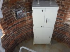 Photograph of curved, internal brick wall of lighthouse. Large white cabinet stands beside wall.  