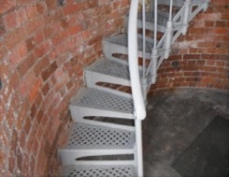 Photograph showing grey lattice spiral stairs descending from upper level, curved around brick tower wall. 