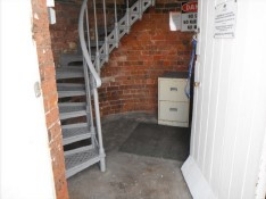 Photograph showing ground floor of lighthouse tower. Grey lattice staircase descends from floor above and wraps around brick tower wall. 