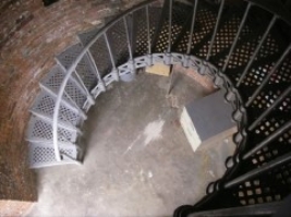 Photograph showing ground floor of lighthouse tower. Grey lattice staircase descends from floor above and wraps around brick tower wall. 