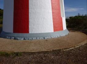 Photograph showing base of red and white striped lighthouse tower. Concrete paving wraps around base of tower.