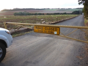 Photograph of road beside a field. A yellow barrier prevents access along the road - sign reads "Road closed Dusk to Dawn"