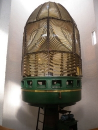 Photograph showing large lighthouse lens with hundreds of prismatic glass pieces arranged in concentric circles. Lens is sitting on a round, green pedestal.