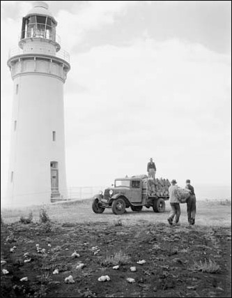 Black and white photograph showing white lighthouse tower. An old car sits beside it, and three men are loading items onto the tray. 