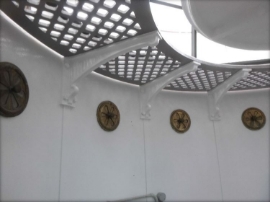 Photograph showing underside of grey, curved lattice floor panels running along edge of lantern room. Four unpainted copper vents are fitted into wall panels.