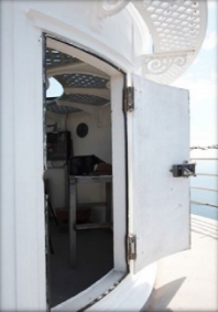 Photograph showing white external lantern room with curved door standing ajar on small lighthouse balcony. 