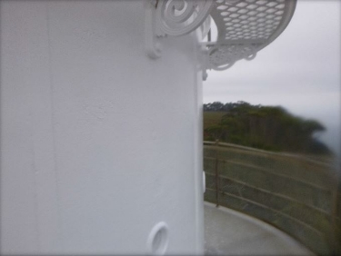 Photograph showing external wall panels of curved, white lantern room. A small balcony wraps around base, and lattice floor panels are fitted above with ornate brackets.