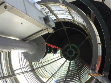 Photograph showing inside of large prismatic lens. A small pedestal with LED light source fitted at top stands in centre of lens. 