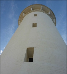 Photograph showing white lighthouse tower against blue sky. Three windows are set deep into the curved walls. 