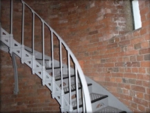 Photograph showing curved, red brick walls. A grey spiral staircase is fixed to the walls and curved up to a higher level. 