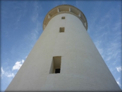 Photograph showing white lighthouse tower against blue sky. Three windows are set deep into the tower walls. 