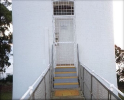 Photograph showing entry door of white lighthouse tower. Three grey steps lead to door. 