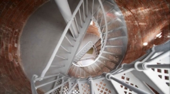 Photograph showing internal view of lighthouse tower. Several grey slate landings fitted inside curved, red brick tower, connected by grey spiral staircase. 