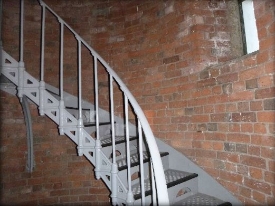 Photograph showing curved, red brick walls. A grey spiral staircase is fixed to the walls and curved up to a higher level. 