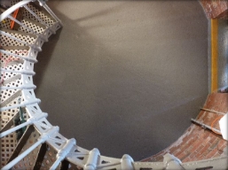 Photograph showing rounded ground floor of lighthouse tower. Grey spiral staircase descends to floor along outer edge. 