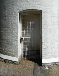 Photograph showing basement door at base of lighthouse tower. Painted steel grille covers door. 