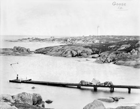 Black and white photo showing a jetty and tramway extending into the sea. A white lighthouse can be seen in the background. 