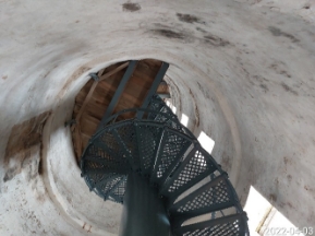 Photograph showing underside of grey spiral staircase fitted within a white tower. Staircase ascends to a timber half-floor above. 