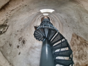 Photograph showing underside of grey spiral staircase fitted within a white tower. Staircase ascends to a timber half-floor above. 