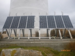 Photograph showing aluminium racks supporting solar panels at base of white lighthouse tower. 