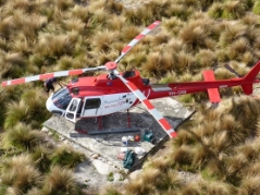 Photograph showing square concrete slab surrounded by grassy vegetation. A red and white helicopter sits on the slab. 