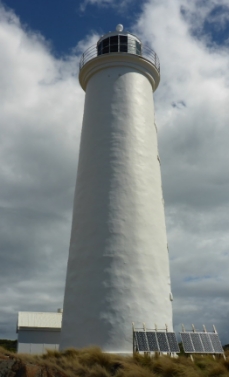 Photograph showing white lighthouse tower against blue, cloudy sky.