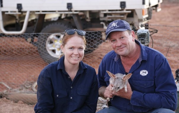 Cassandra Arkinstall and Save the Bilby CEO Kevin Bradley crouch holding a bilby in the outback.