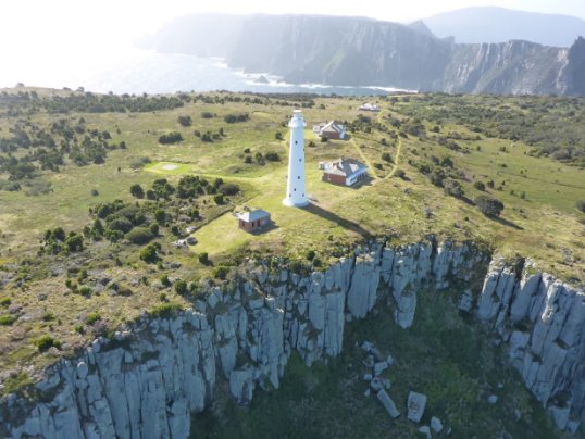 Photograph showing tall, white lighthouse at edge of rocky cliffs. Several buildings are scattered around the green cliff top, and cliffy promontories can be seen in the distance behind the tower.