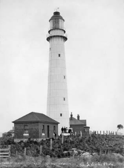 A black and white photograph showing a tall, white lighthouse tower between two brick buildings. A collection of people stand at the base of the tower. 