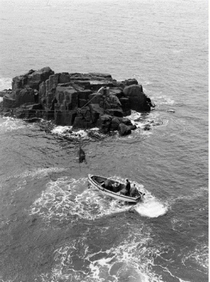 Black and white photograph showing flying fox pulley system anchored to rock, slinging cargo along. A boat with a small number of people sits in the water below.  