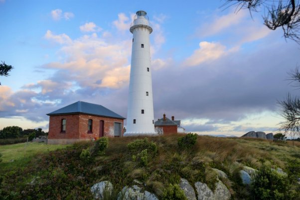 Photograph showing tall, white lighthouse tower beside two small brick buildings. 