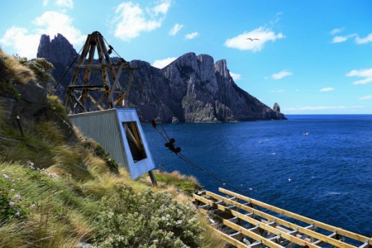 Photograph showing winch shed set on edge of cliff with pulley system attached. Another cliffy island rises out of ocean in background. 