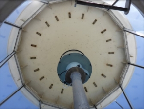 Photograph showing ceiling of round, glazed lighthouse lantern room with steel column fitted with beacon. 