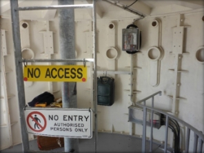 Photograph showing inside a lighthouse room. Beside a steel column spanning the height of the room, a ladder is fitted inside with 'No access' and 'No entry' signs. 