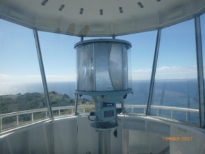 Photograph showing beacon sitting atop a steel post within a small, glazed lantern room. 