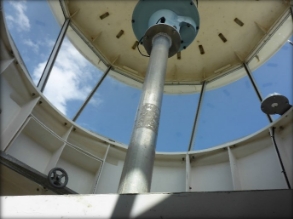 Photograph showing ceiling of round, glazed lighthouse lantern room with steel column fitted with beacon. 