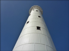 Photograph showing tall, white lighthouse tower consisting of cast iron panels bolted together. Several windows are fitted into the tower at varying heights. 